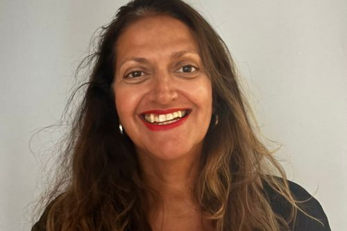 Smiling woman with long brown hair, red lipstick, and hoop earrings against a plain background
